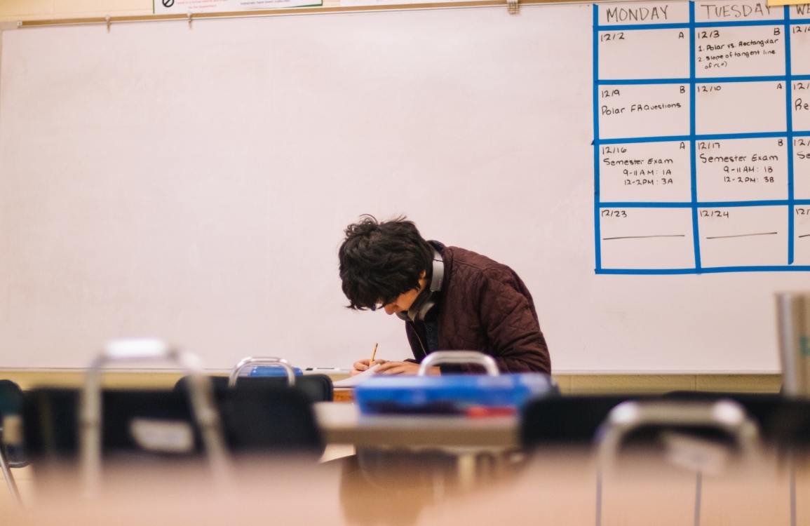 A person sits in an empty classroom with headphones around their neck as they work. Partial calendar of the month is drawn on the dry eraser board behind them.